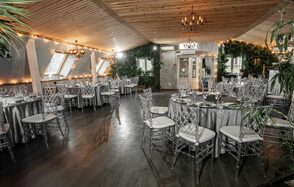 Loft - bar under closure in Peterhof. Банкетный зал с наклонным деревянным потолком и деревянным полом, круглые столы покрыты серыми скатертями, вокруг стулья с прозрачными спинками. На стенах окна и зелень. Освещение от люстр и гирлянд.
