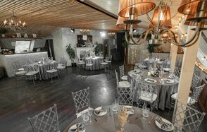 Loft - bar under closure in Peterhof. Банкетный зал с круглыми столами, накрытыми серыми скатертями. Прозрачные стулья вокруг столов. В центре лампы с коричневыми абажурами. Барная стойка слева с деревянными элементами. Декор в виде растений и тёплая светодиодная гирлянда на стене.