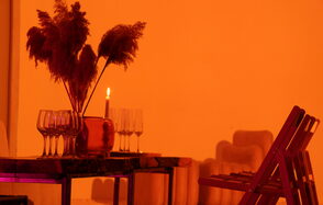 Светлый лофт со светомузыкой и барной стойкой. Пространство оформлено в теплых оранжевых тонах. Стол с вазой, наполненной сухими растениями и зажжённой свечой. Несколько бокалов на столе. Рядом располагается стул. В интерьере видны мягкие изгибы мебели.