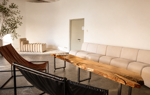  Light loft with light music and a bar. Просторная комната с белыми стенами и бетонным полом. Большой светлый диван у стены, бежевое кресло на ковре, два темных стула. Деревянный стол на металлическом каркасе, дверь без декора. Натуральное освещение. Угловое растение.