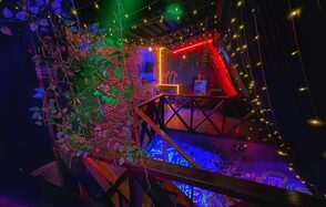 Лофт-бар. Крытое пространство с кирпичными стенами, балконом с деревянными перилами и растениями. Яркое разноцветное освещение, гирлянды на потолке. Обстановка с барной стойкой, стульями, квадратным столиком и черными занавесками.