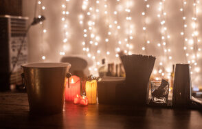 Большой исторический лофт. Барная стойка с ведром для льда, свечами, стаканами и трубочками. На заднем плане светодиодные гирлянды создают мягкое освещение. Стол выполнен из темного дерева. Цвета: теплые оттенки, преобладают желтый и оранжевый.