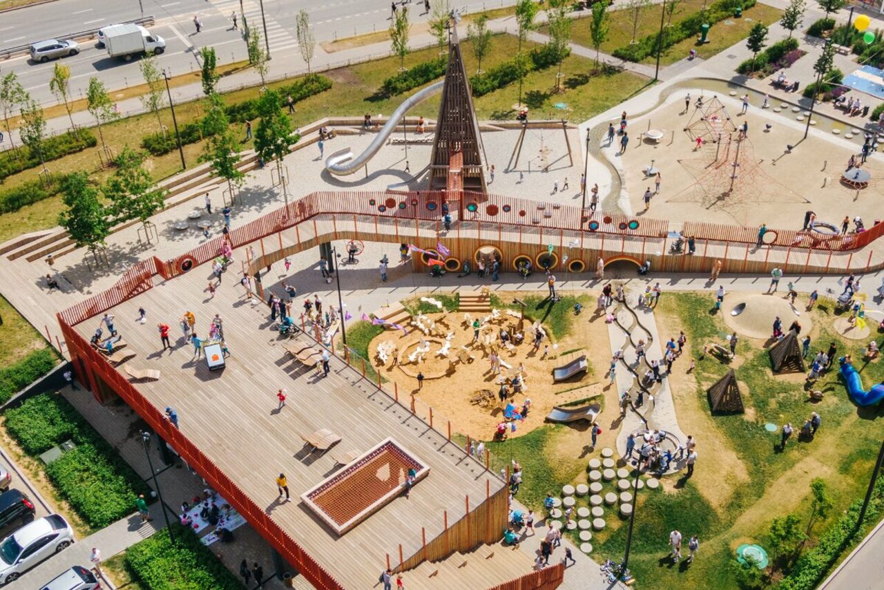 Арендовать Площадка в Москве c одной фотографией. Детская площадка с несколькими уровнями; деревянное покрытие, металлические горки, песочница, сетки для лазания, скалодром, зелёные участки; ограждение из дерева и металла; много посетителей.